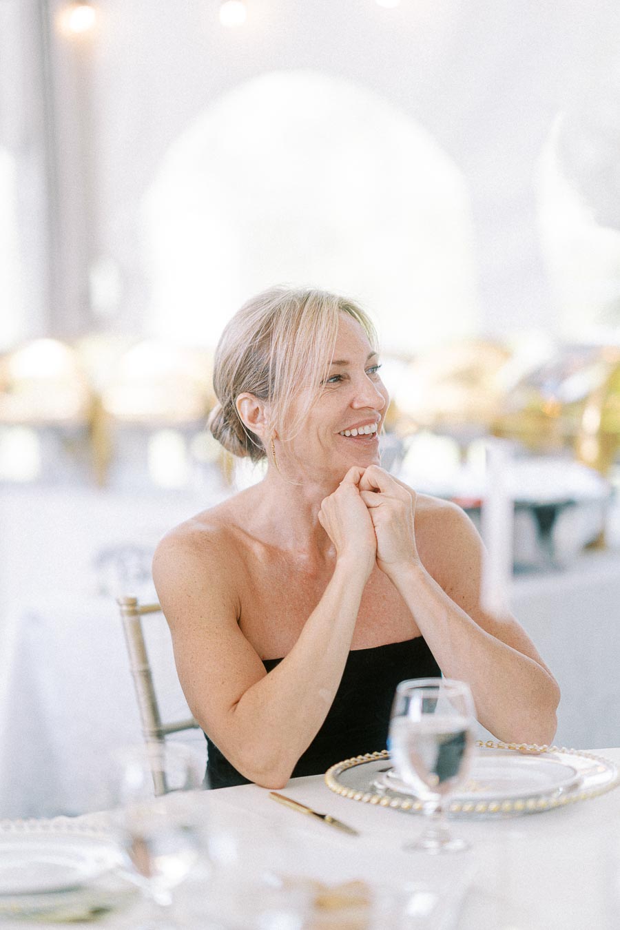 Smiling woman sitting at an elegantly set dining table, clasping hands in a joyful atmosphere.