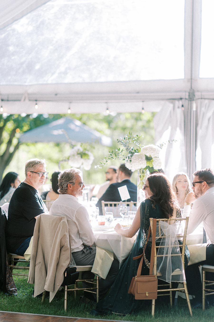 A group of elegantly dressed guests sit at a beautifully decorated table under a transparent tent during a sunny outdoor event.