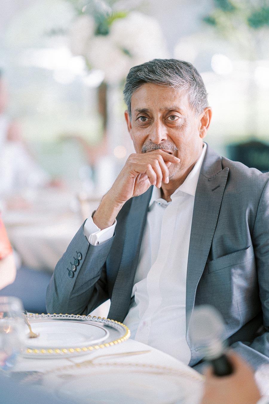 Elegant man in a grey suit sitting at a table, thoughtfully holding his chin, with blurred background featuring a bright, airy setting and floral decorations.