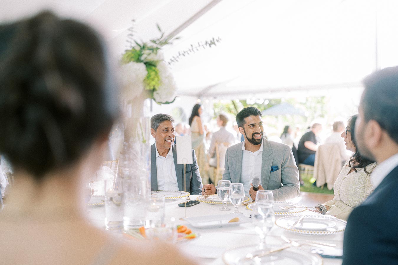 A group of elegantly dressed people sitting around a table at an outdoor wedding reception, engaging in conversation and surrounded by beautifully arranged flowers and glassware.