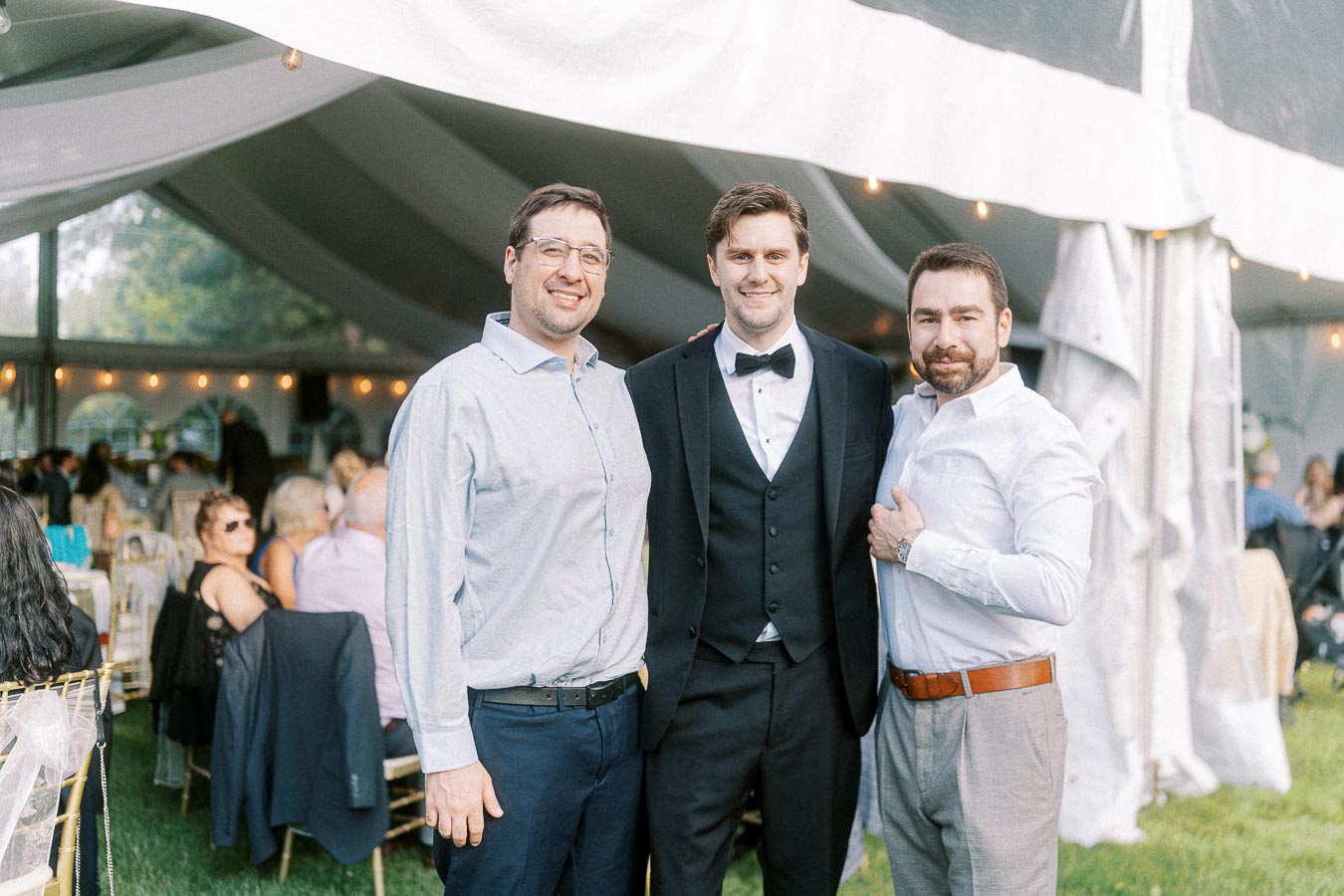 Three men posing together under a decorated tent at a formal outdoor event, with guests seated in the background. One man is dressed in a tuxedo, and the others are wearing dress shirts, creating a celebratory atmosphere.
