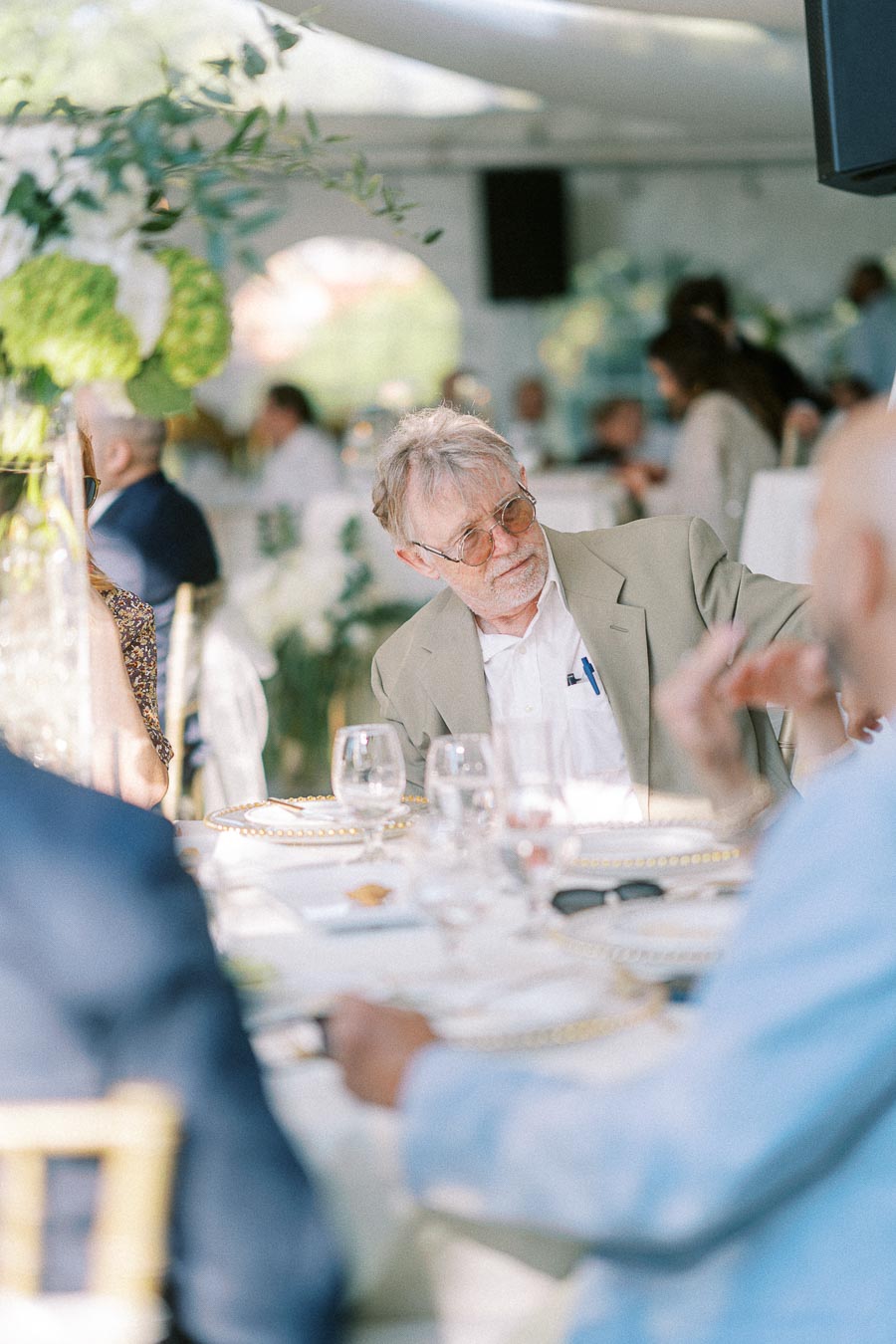 An elderly man in a light-colored jacket and glasses engaging in conversation at a formal outdoor gathering, surrounded by elegantly set tables and blurred guests in the background.