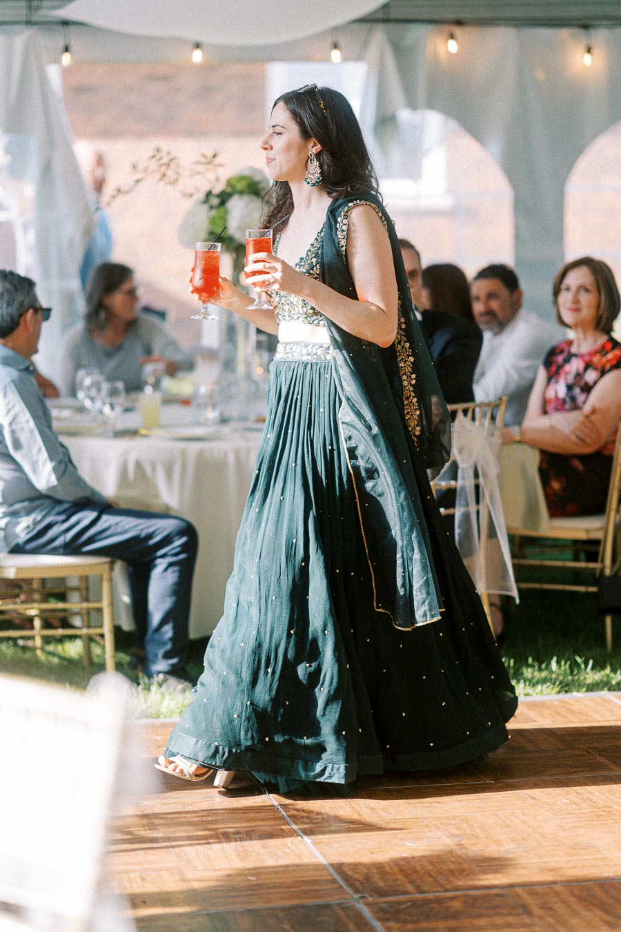 Woman in an elegant green lehenga carries red drinks at an outdoor event.