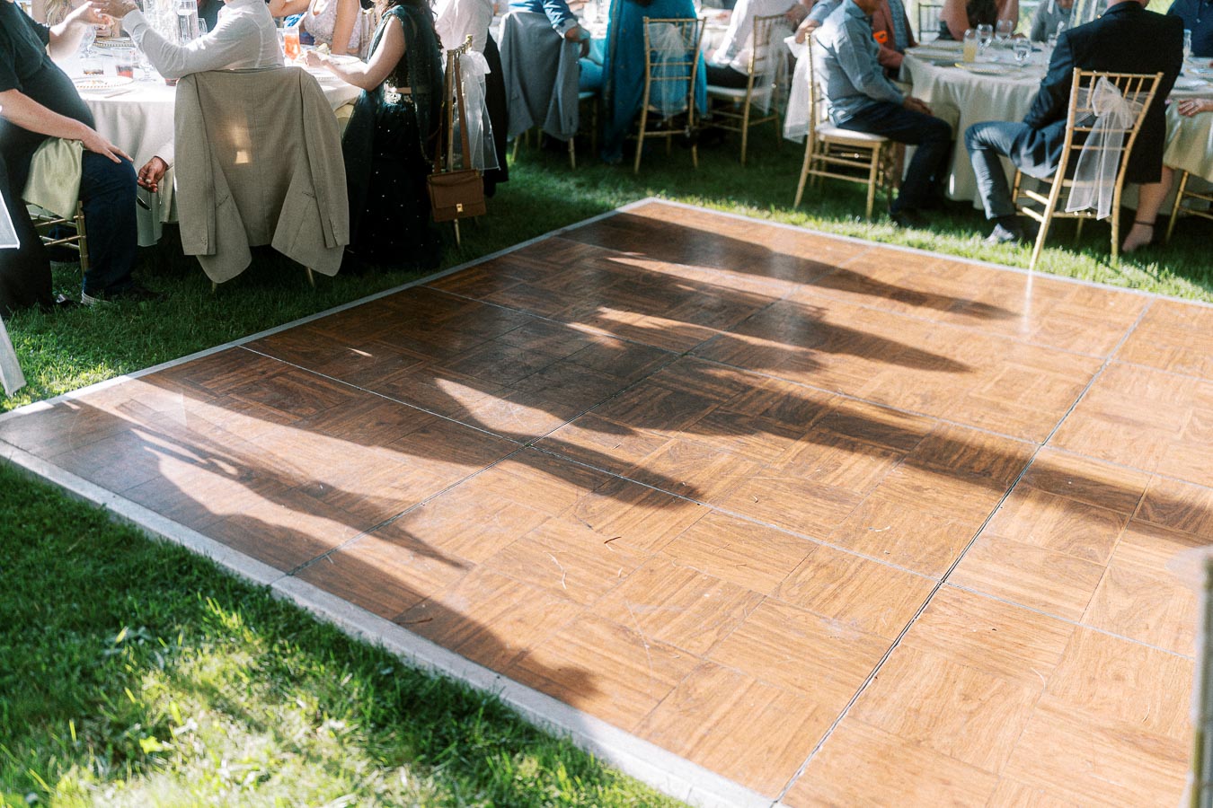 Outdoor wedding reception with guests sitting at tables and sunlight casting shadows on a wooden dance floor, surrounded by greenery.