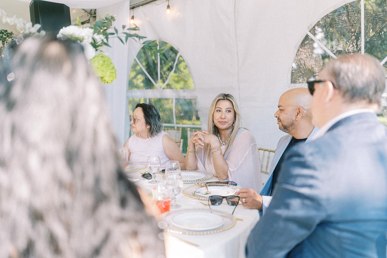 Group of people sitting at an elegantly set table during an outdoor event, with clear sky and greenery visible through tent windows; they are engaged in conversation, creating a warm, social atmosphere.