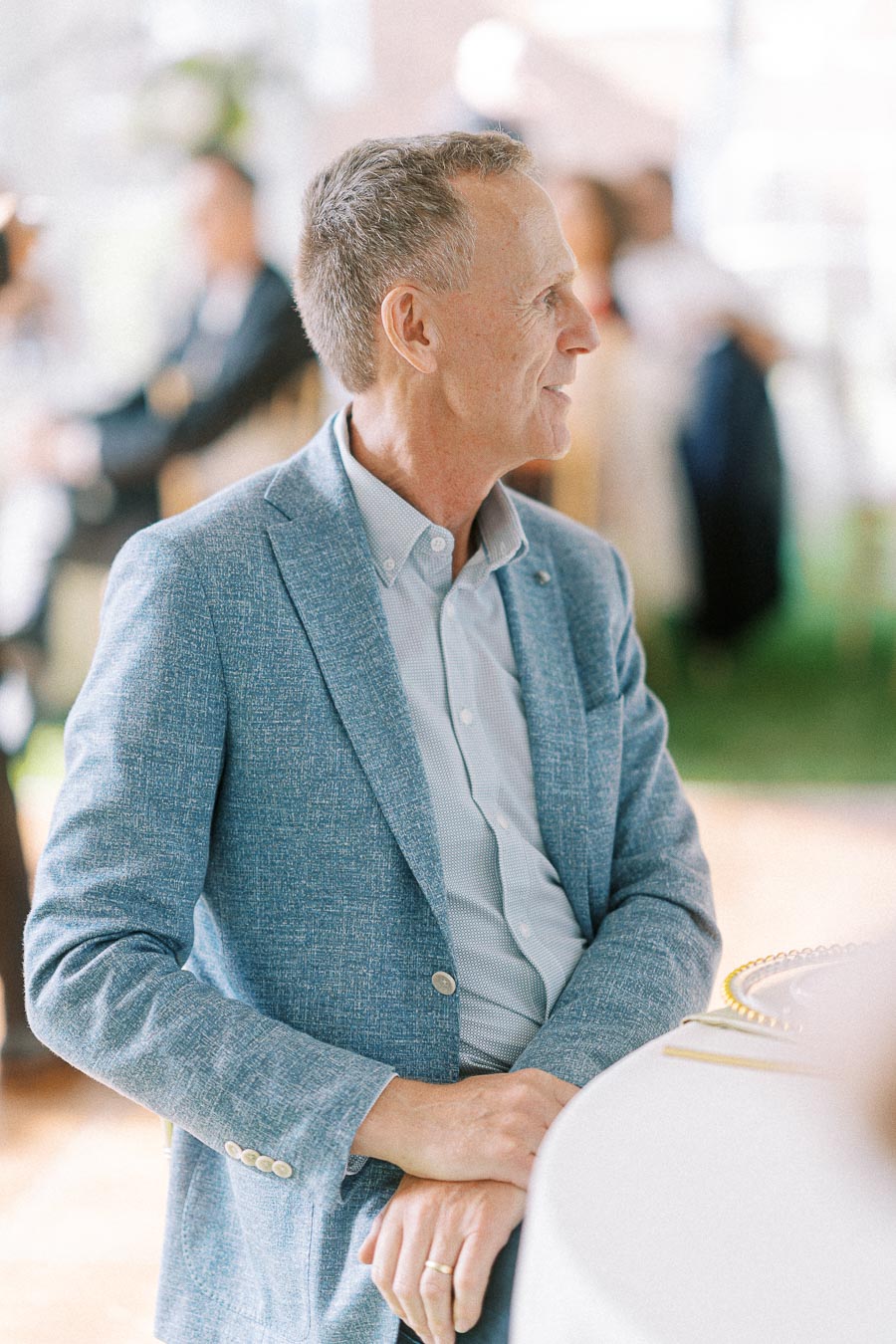 Elderly man in a stylish light blue blazer sitting at a formal event table, looking engaged and thoughtful, with blurred background guests at a social gathering.