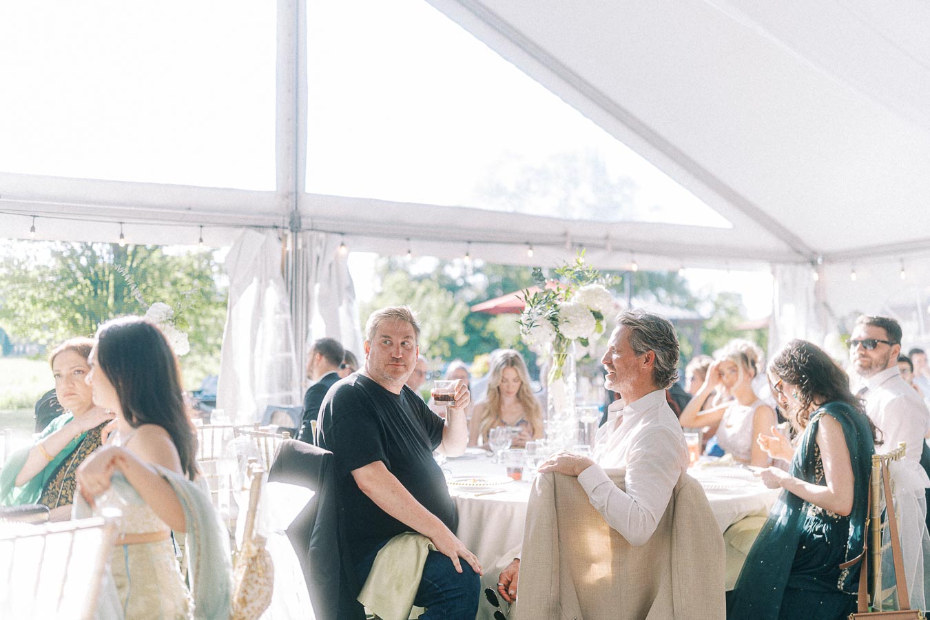Guests enjoying a sunny afternoon at an outdoor wedding reception under a tent, featuring elegantly decorated tables with floral centerpieces and a vibrant social atmosphere.
