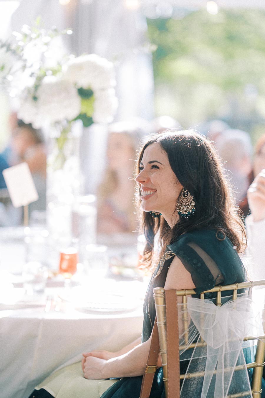 Smiling woman in elegant attire at a sunlit outdoor event, seated on a gold chair with floral centerpiece in the background.