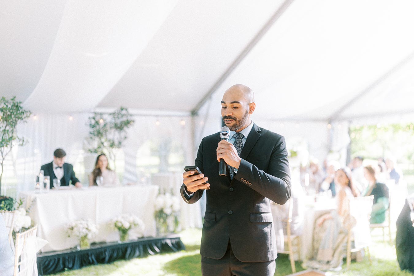 A man in a suit delivers a speech using a microphone at an outdoor wedding reception under a white tent, with elegantly dressed guests seated at tables and a decorated table in the background.