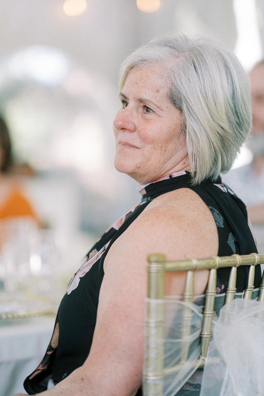Elderly woman with short gray hair, seated on a decorative chair, wearing a floral black top at a formal event.