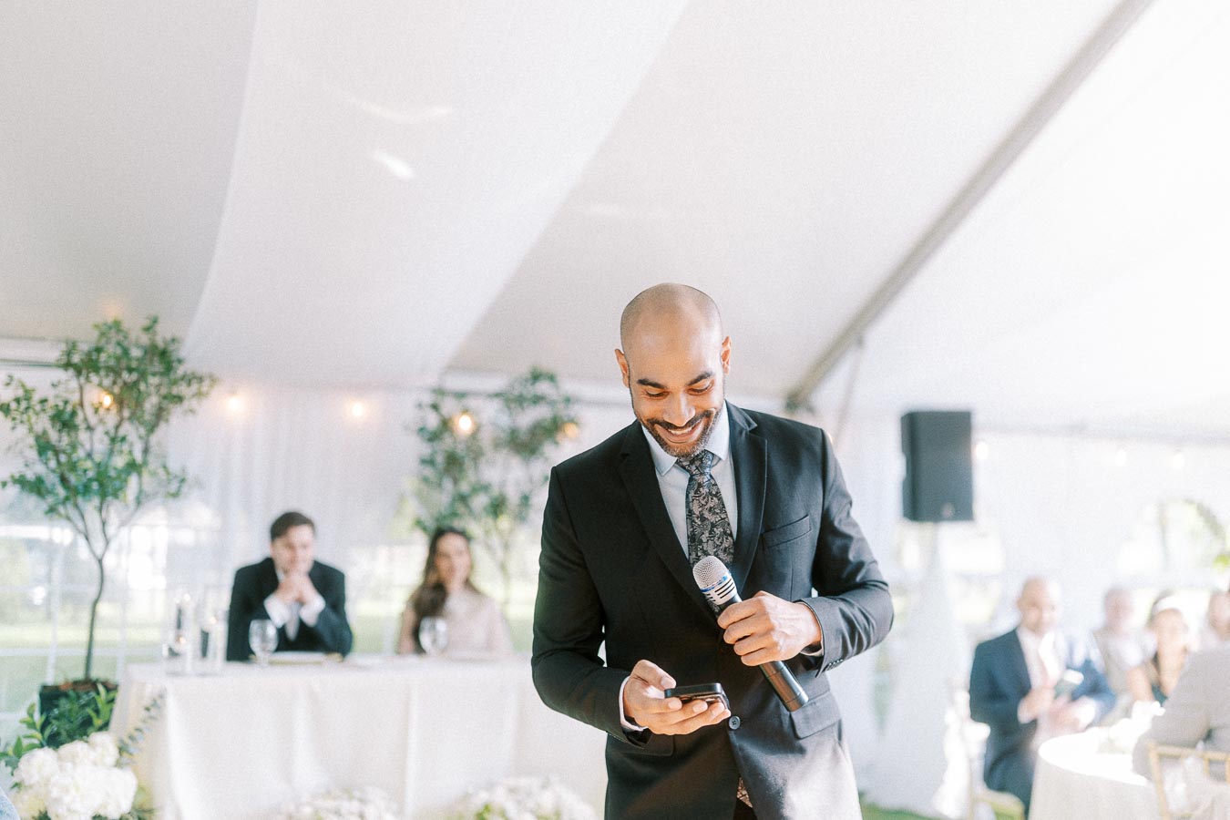 A man in a suit smiling and holding a microphone, preparing to give a speech at a wedding reception inside a tent, with guests seated in the background.