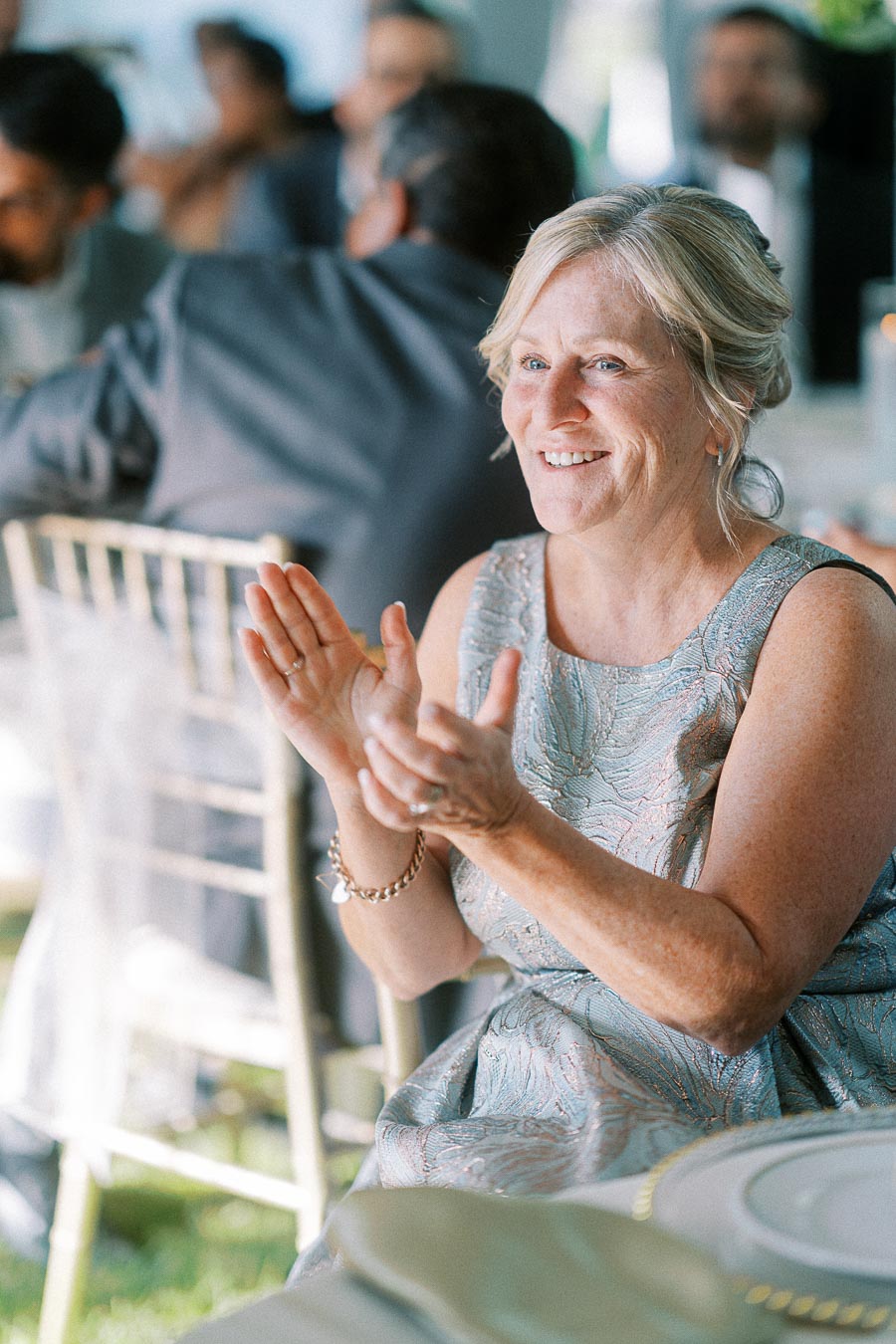 Elderly woman at a wedding reception, smiling and clapping, wearing a silver dress, with blurred guests in the background.