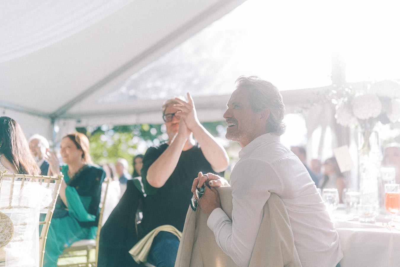 Guests enjoy a light-filled tented event, with a smiling man in the foreground holding his jacket and sunglasses, as others applaud in the background.