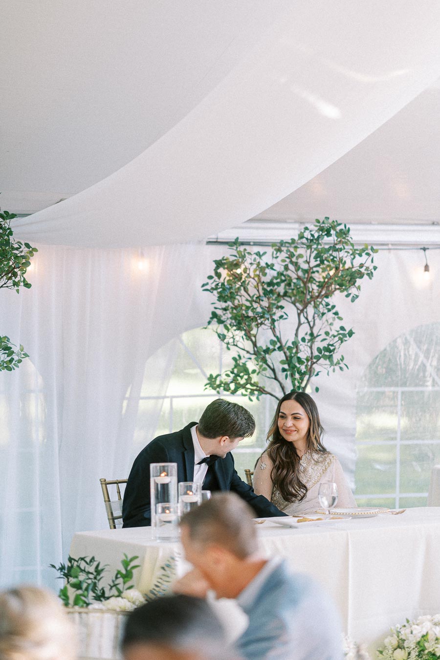 Bride and groom sitting at a decorated table under a white canopy during a wedding reception, surrounded by greenery and candles.