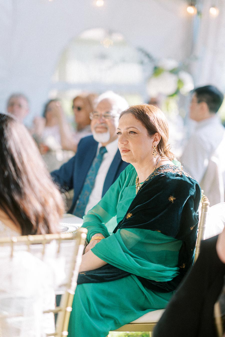 Elegant woman in a green traditional outfit attending an outdoor event, surrounded by other seated guests under a tent during daylight.