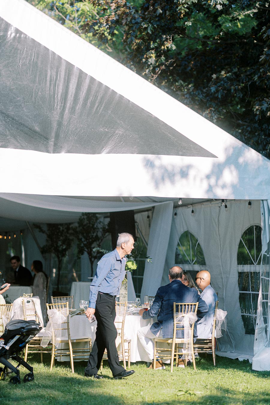 Outdoor wedding reception under a white tent with guests seated at elegantly decorated tables, surrounded by greenery and natural light.