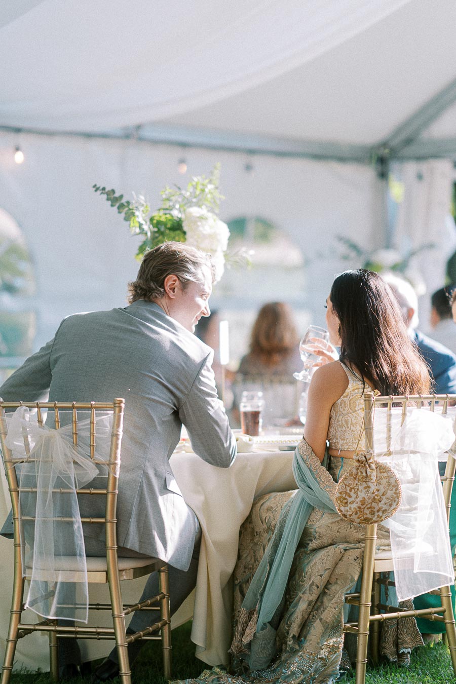 Elegant outdoor wedding reception with a couple seated at a beautifully decorated table in a white tent. The man is wearing a light gray suit, while the woman is in an embellished traditional dress with a sheer shawl, both enjoying the refined ambiance and natural daylight filtering through the space.