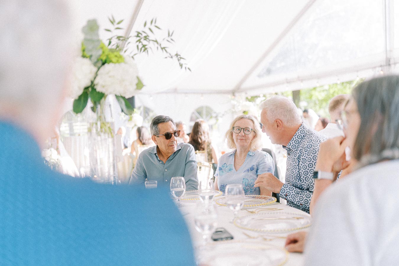 Group of people enjoying a conversation at an elegant outdoor party, seated around a decorated table under a white canopy with floral arrangements.