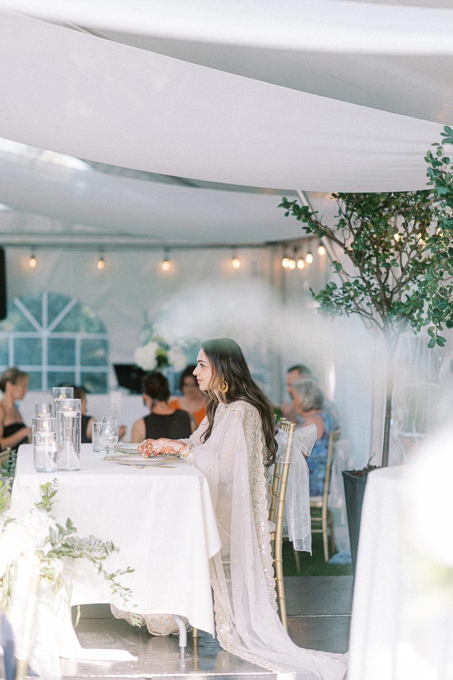 Elegant woman in a white saree sitting at a beautifully decorated dining table in a well-lit tented event setting, surrounded by guests and adorned with candlelit vases and floral arrangements.