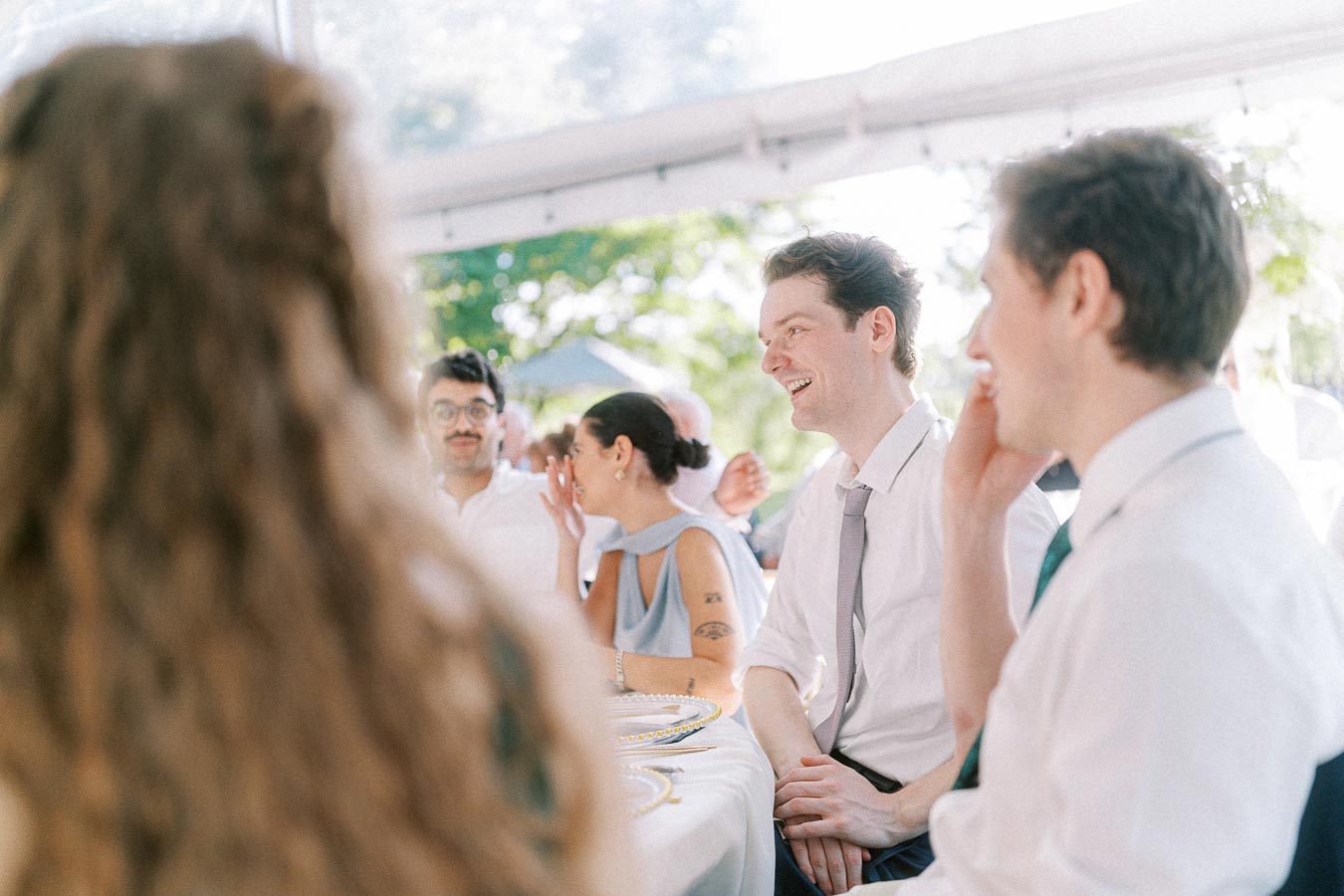 Group of people smiling and chatting at an outdoor event under a canopy, with natural sunlight filtering through trees in the background.