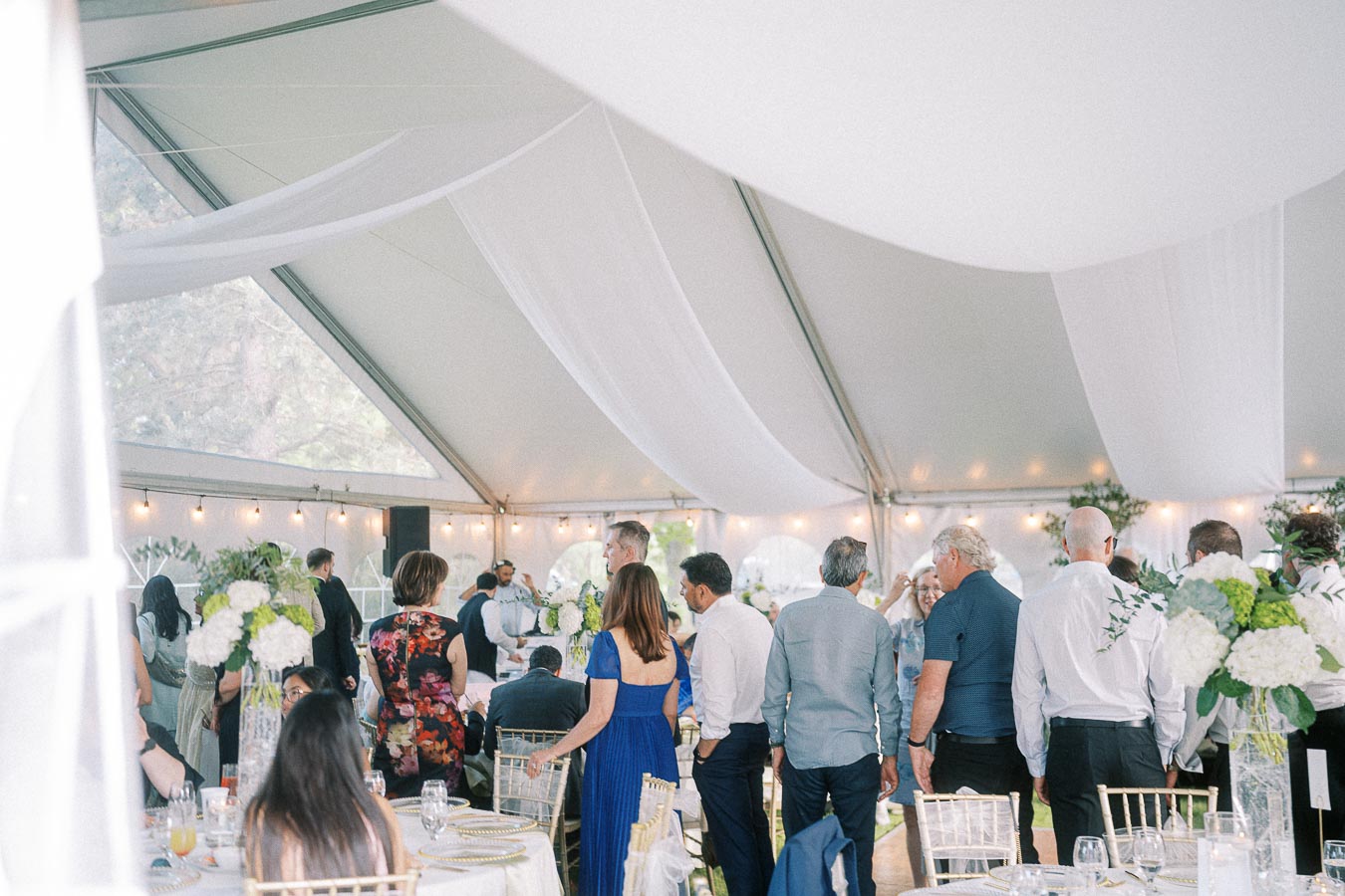 Elegant outdoor wedding reception under a beautifully decorated white tent, featuring guests mingling and exquisite floral centerpieces in a festive, atmospheric setting.