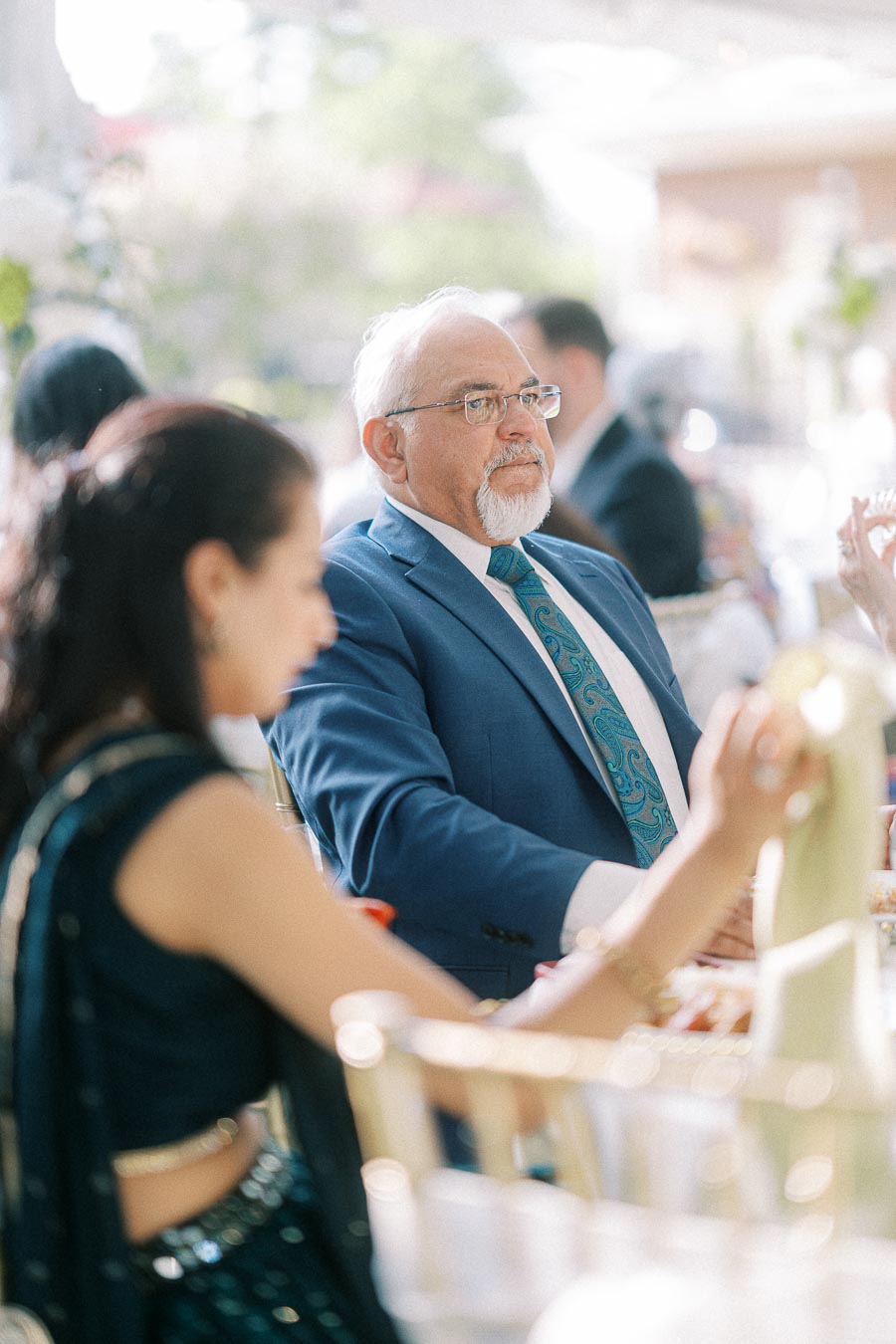 Elderly gentleman in a blue suit and glasses attentively listening at an elegant indoor event, with a blurred background showing other attendees.