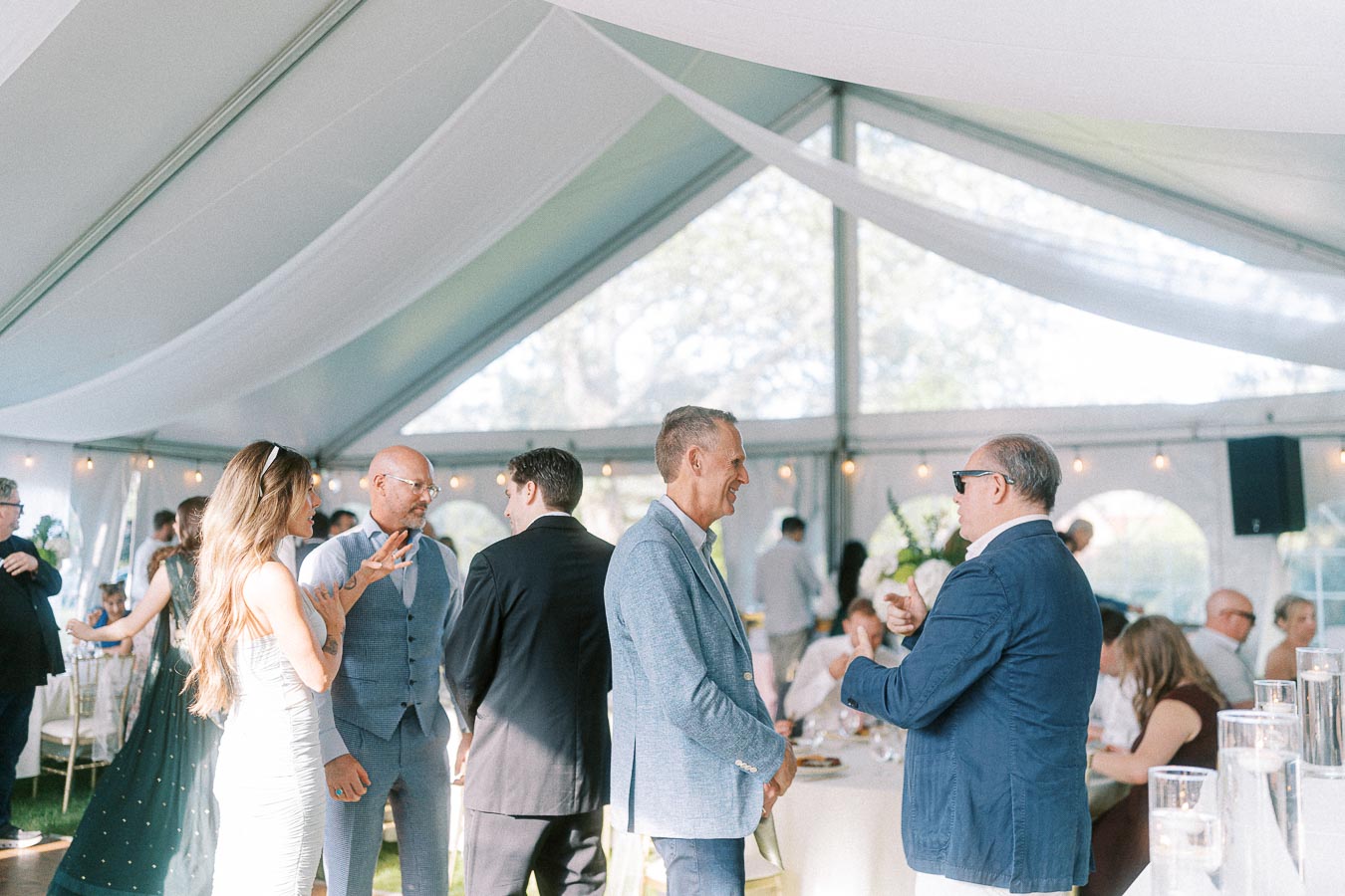 People socializing at an elegant party under a white marquee tent, with soft natural light and ambient decor, including string lights and greenery, creating a sophisticated and festive atmosphere.