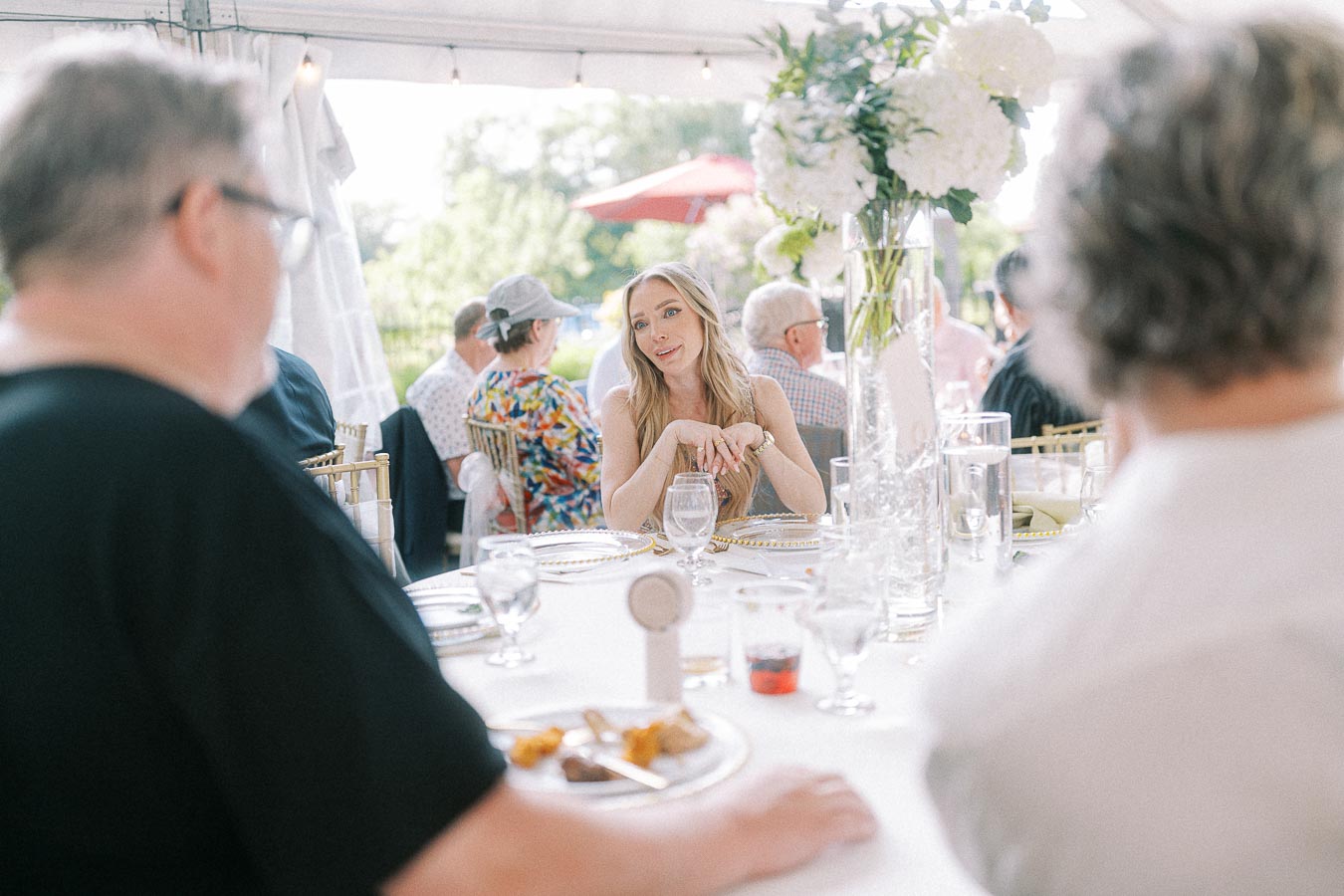 A woman engaging in conversation at a beautifully decorated outdoor reception, surrounded by elegantly dressed guests and floral centerpieces, under a white canopy.