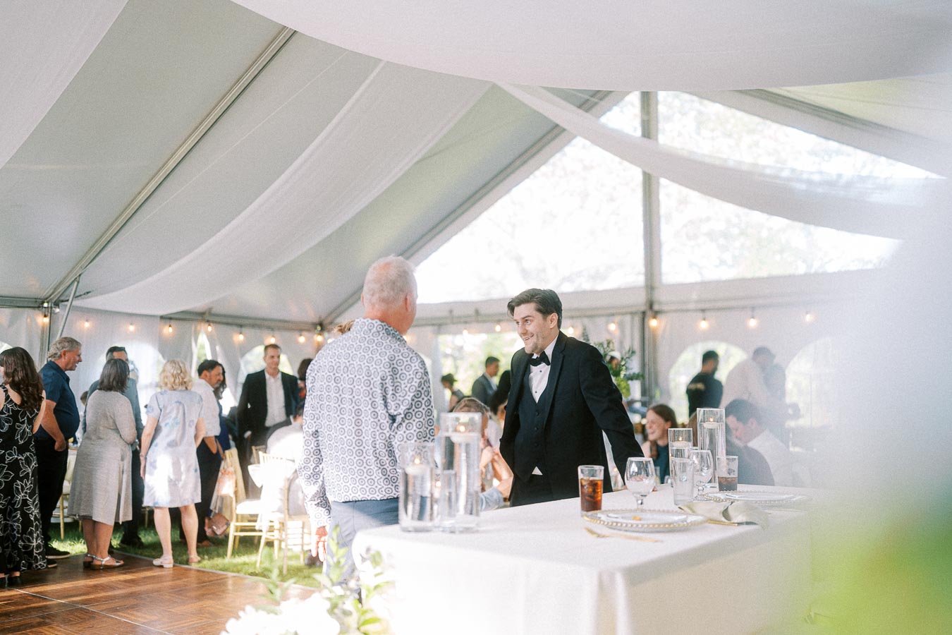 Guests mingling at a wedding reception inside a beautifully decorated tent, featuring a bartender in formal attire, elegant table settings, and soft ambient lighting.