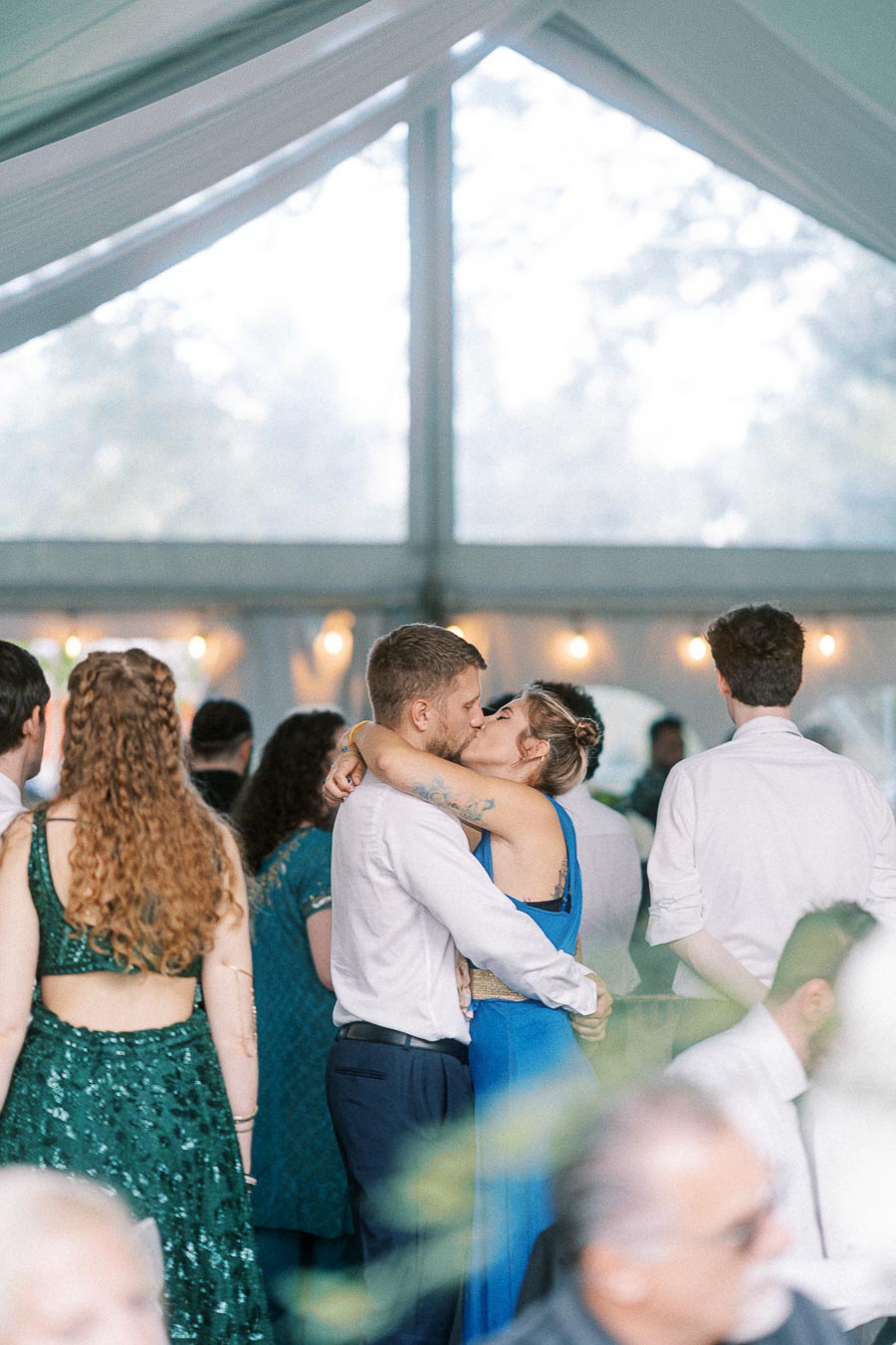 A couple embracing and kissing at a wedding reception, surrounded by guests in a beautifully decorated marquee with fairy lights.