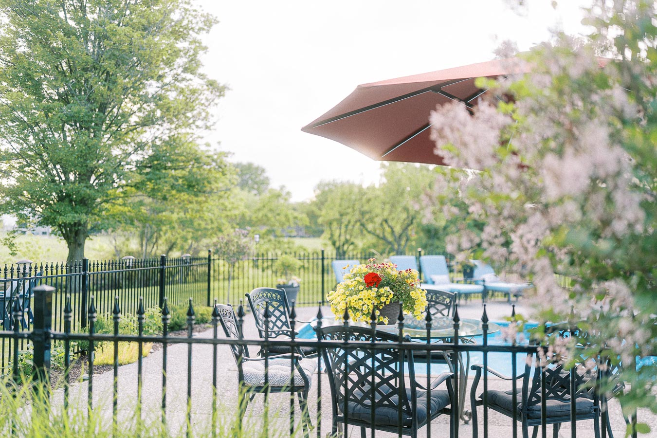 Outdoor patio with black wrought iron furniture, a large red umbrella, a vibrant flower arrangement, and a peaceful view of trees and a pool in the background.