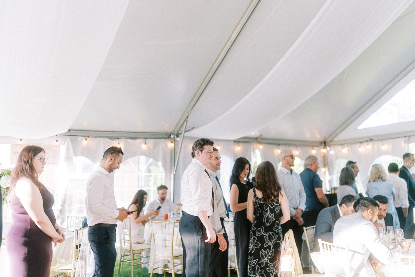 Guests mingling at a sunlit wedding reception under a white tent with elegant decor and string lights.