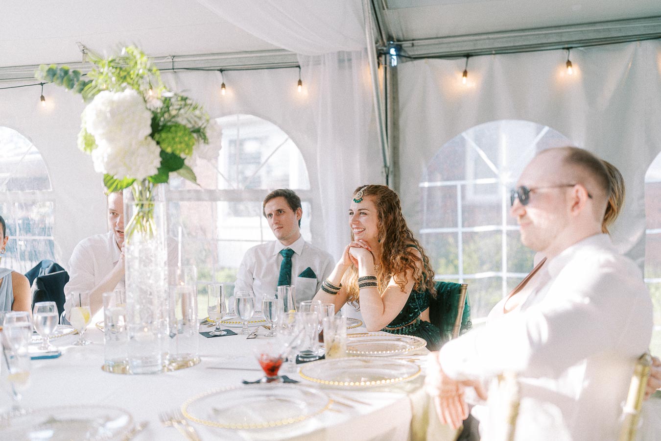 Guests enjoying a formal event seated at an elegantly set table with white tablecloth, delicate glassware, and a centerpiece of lush flowers in a sunlit tented venue.
