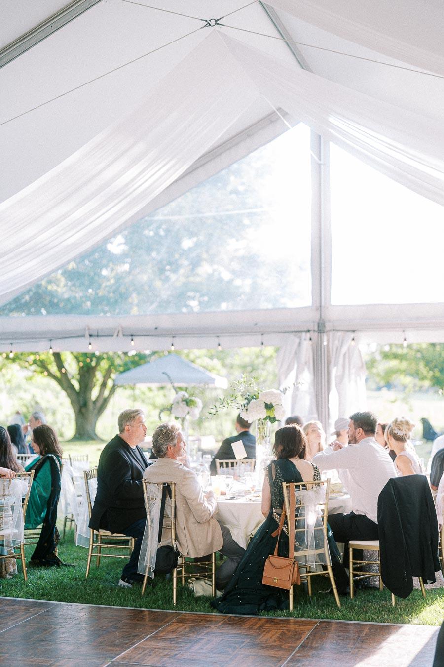 Elegant outdoor wedding reception with guests seated at a round table under a white canopy, surrounded by greenery and soft natural light.