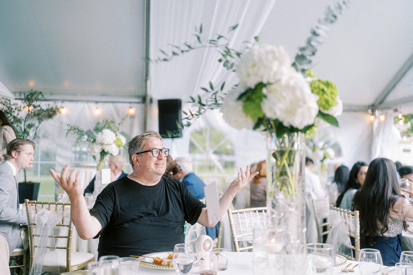 Man smiling and gesturing with hands at a formal event under a tent, surrounded by elegantly decorated tables with floral centerpieces, natural lighting, and seated guests in background.