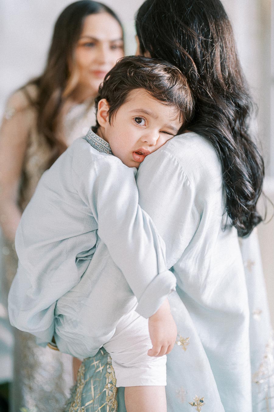 A young child in a light blue traditional outfit being carried by a woman with long dark hair. The child appears to be winking and resting his head on her shoulder, while another woman is softly blurred in the background.