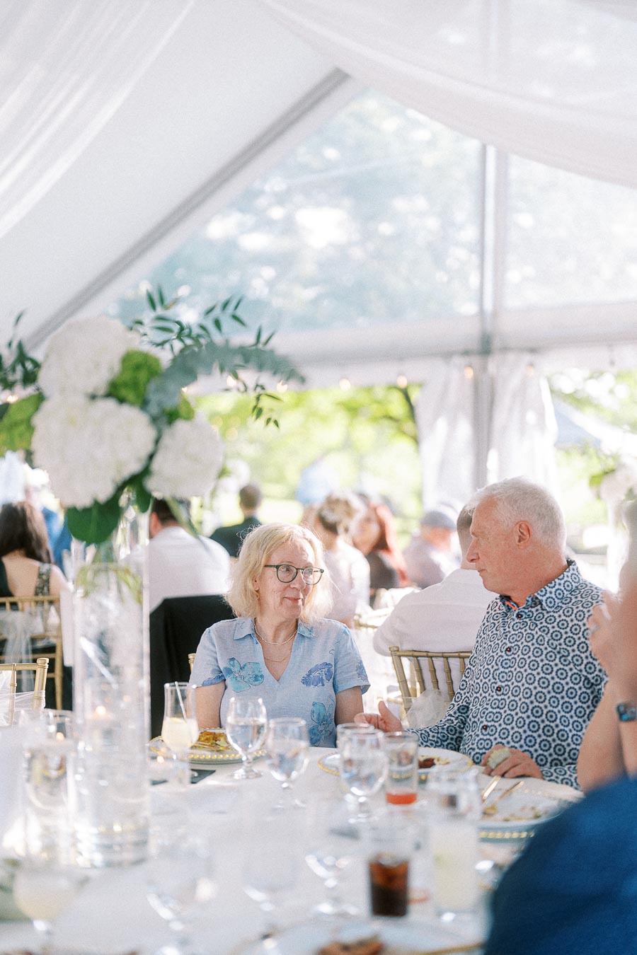 Elderly couple enjoying a conversation at a beautifully set table during an outdoor wedding reception under a white tent.