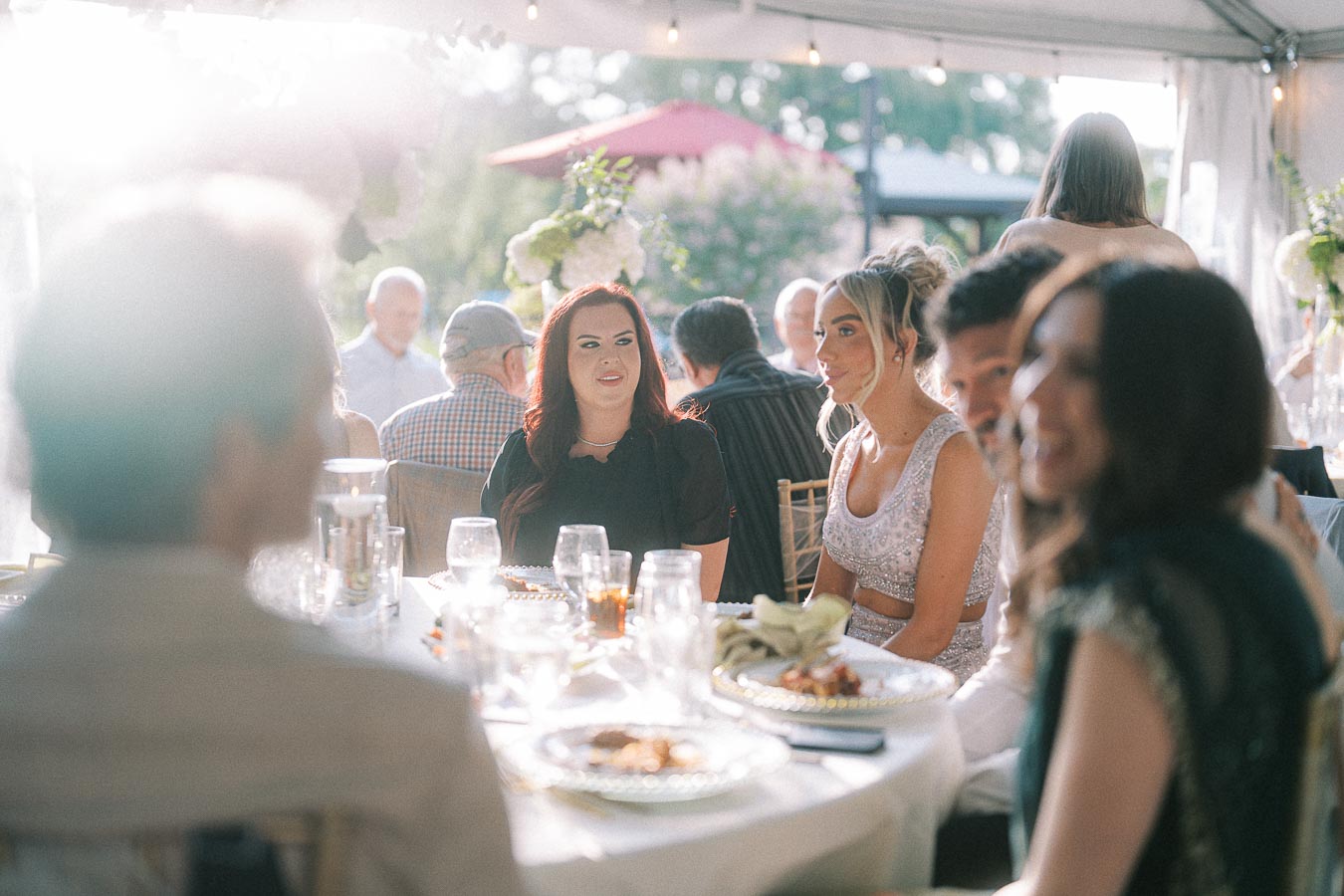 Group of people enjoying a sunny outdoor event under a tent, seated at a table with plates of food, surrounded by greenery.