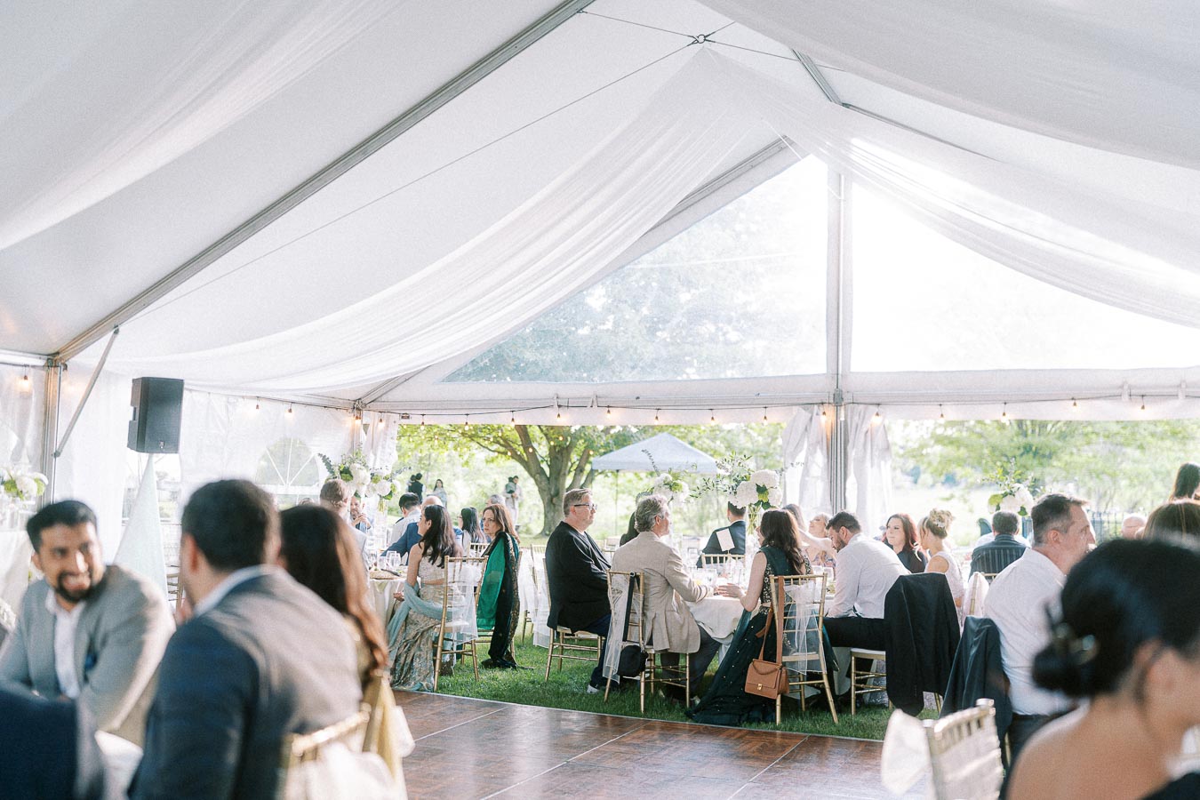 Elegant outdoor wedding reception under a white tent with guests seated at round tables, surrounded by natural greenery and soft lighting.