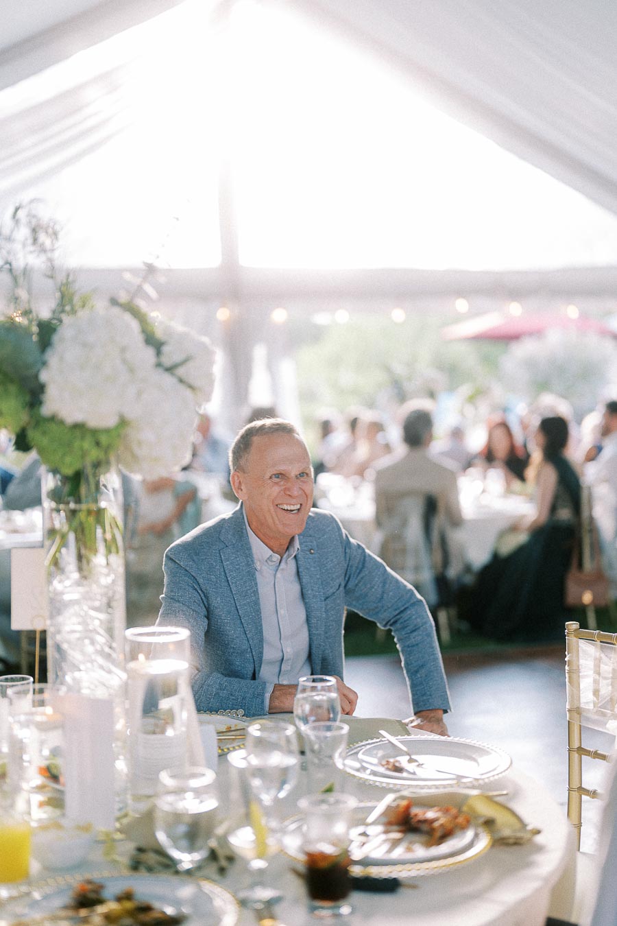 A well-dressed man smiling at a formal outdoor event, seated at a table adorned with elegant dinnerware, surrounded by guests under a white canopy with sunlight filtering through.