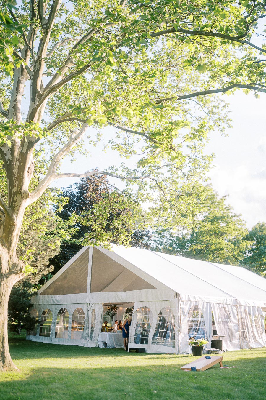 Outdoor wedding reception in a sunlit garden with a large white event tent under a lush green tree.