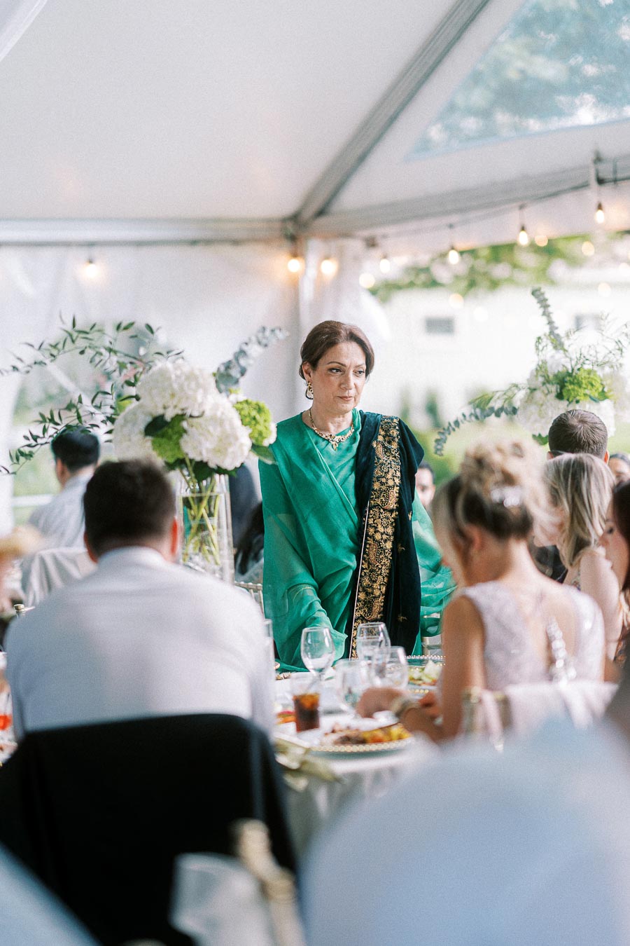 Elegant woman in a green sari standing at a beautifully decorated wedding reception table under a tent, surrounded by seated guests and white floral arrangements.