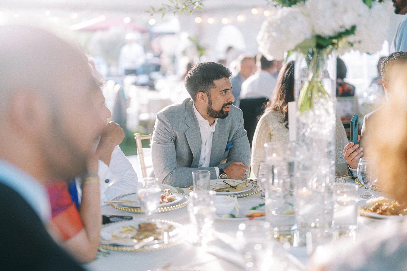 Guests enjoying a formal outdoor event, seated around an elegantly set table adorned with a white floral centerpiece, engaged in conversation and dressed in formal attire.