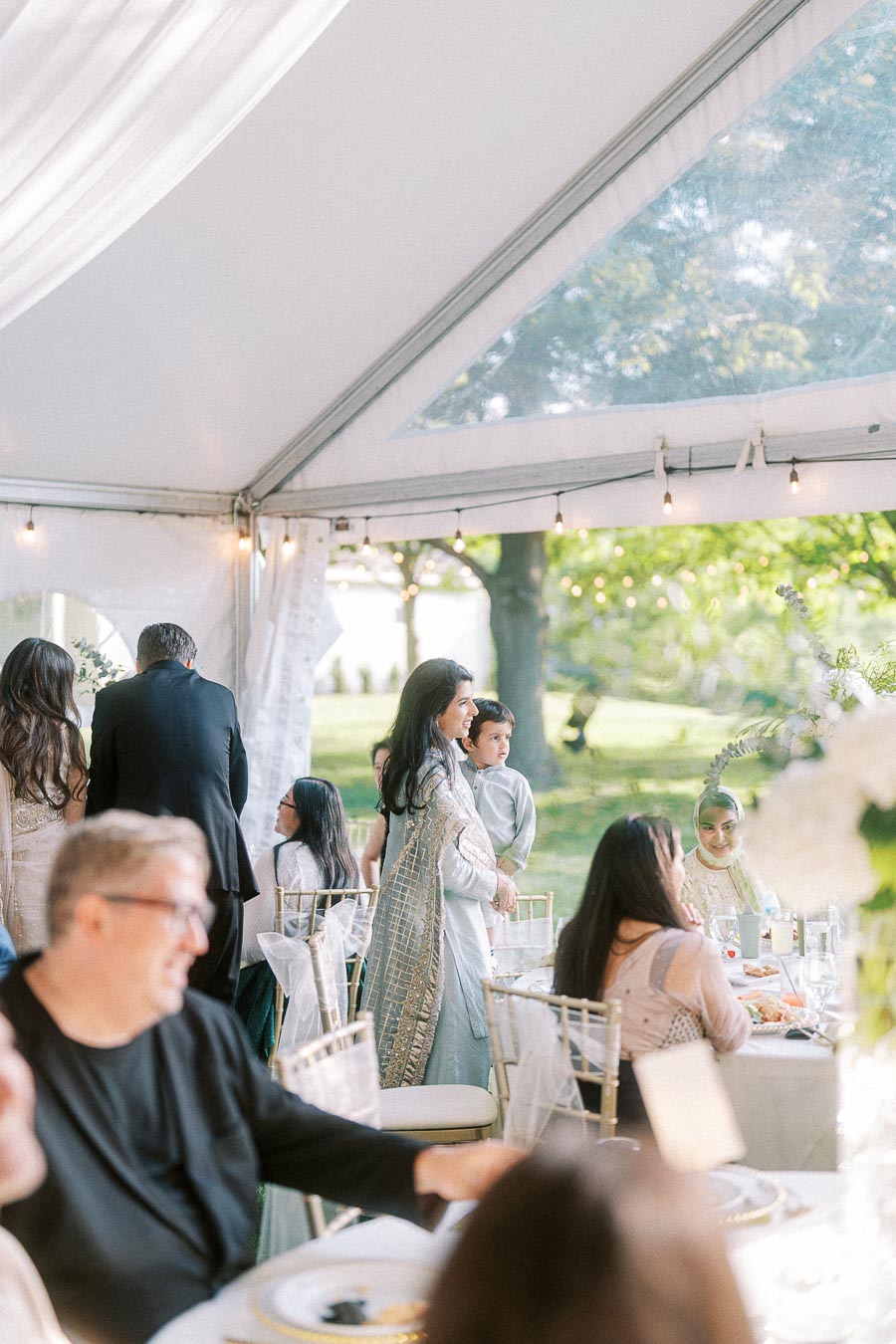 Elegant outdoor wedding reception under a tent with guests in traditional attire, soft lighting, and scenic greenery in the background.