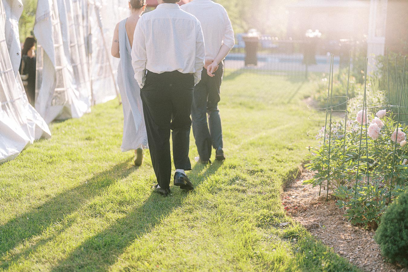 A group of people walking on a sunlit grassy path next to a blooming garden during an outdoor event.