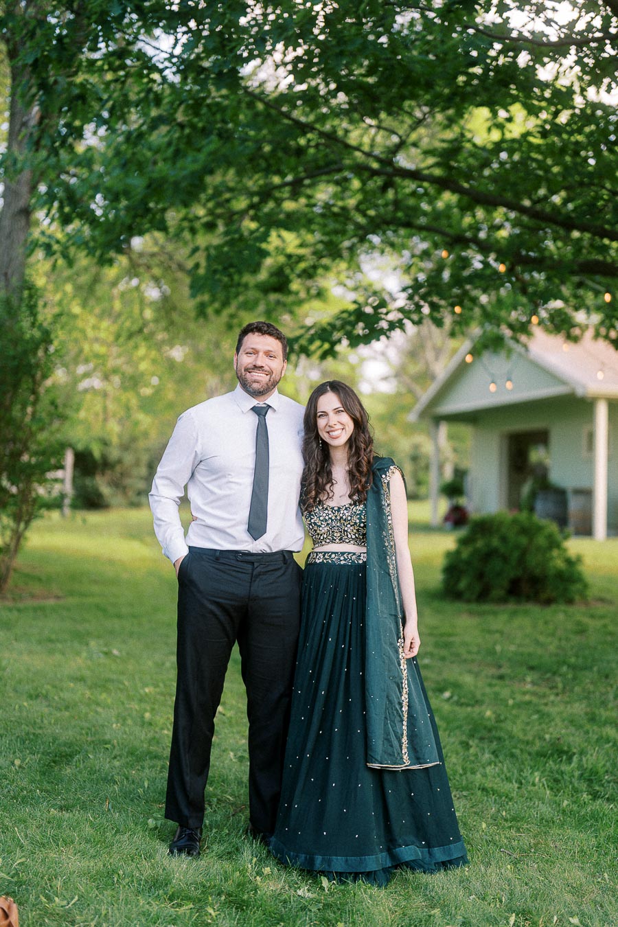 A couple standing in a lush green garden, with the man dressed in a formal white shirt and black tie, and the woman wearing a traditional, elegant dark green dress, smiling under the shade of a large tree.