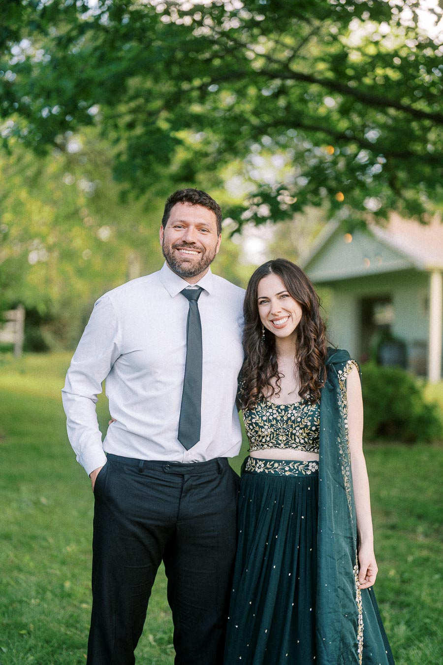 A happy couple posing outdoors in formal attire, with the man in a white shirt and black tie and the woman in a dark green embellished dress. They are surrounded by greenery and trees, with soft sunlight filtering through the leaves.
