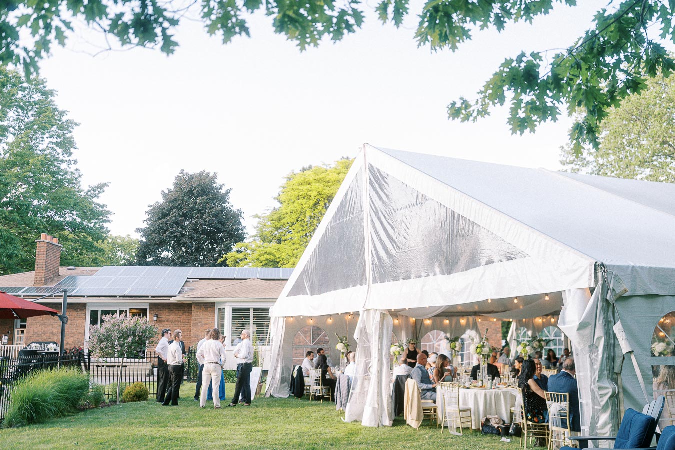 Outdoor event with guests gathered under a large white marquee tent, featuring elegant table settings and floral arrangements, surrounded by lush greenery and a building with solar panels in the background.