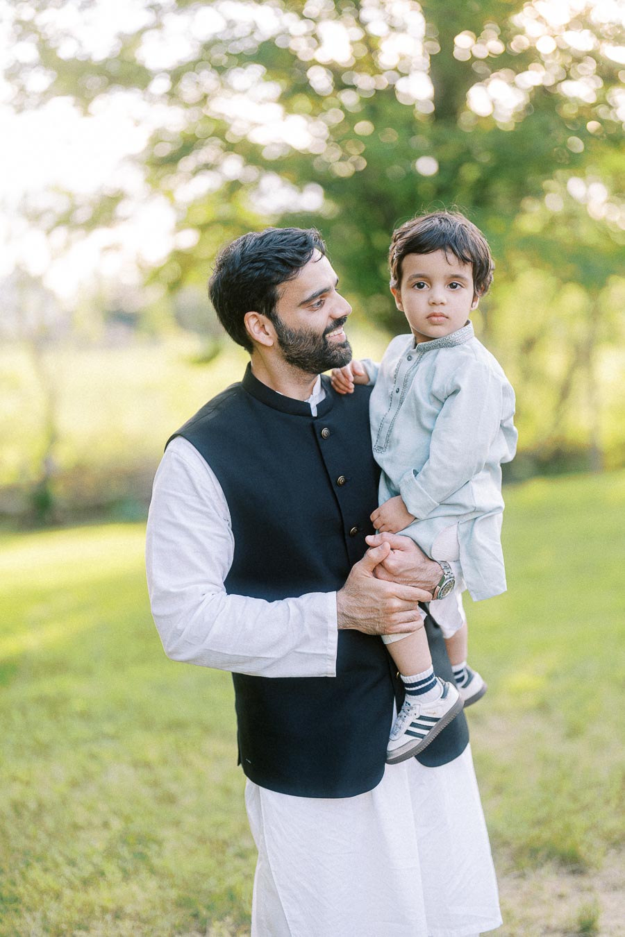 Father holding son outdoors in traditional attire, enjoying a sunny day in a green park.