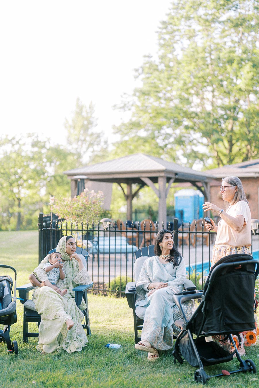 A group of women and a child sitting on chairs in a sunny outdoor garden, next to a stroller and a gazebo in the background.
